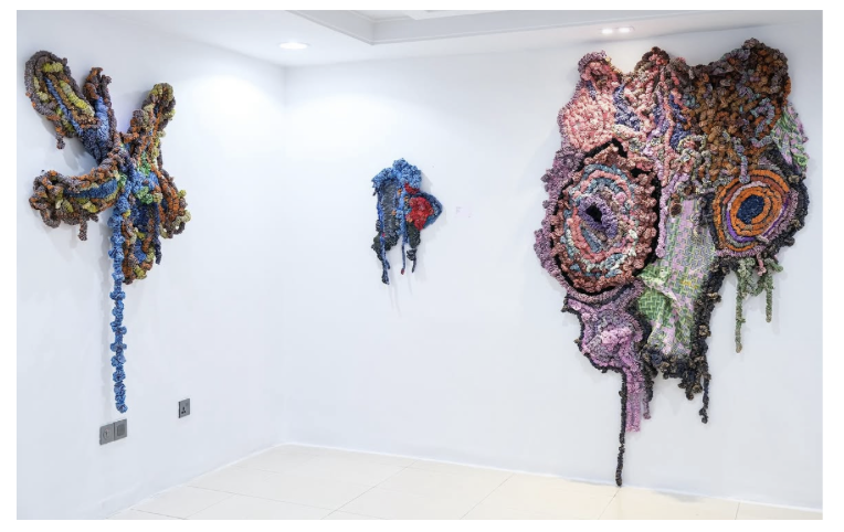 Textile wall art exhibit with textured fabric formations. The left piece features blue and orange hues, the middle presents blue, grey and red tones, and the right showcases pinks, purples, and greens, exuding a tactile presence.